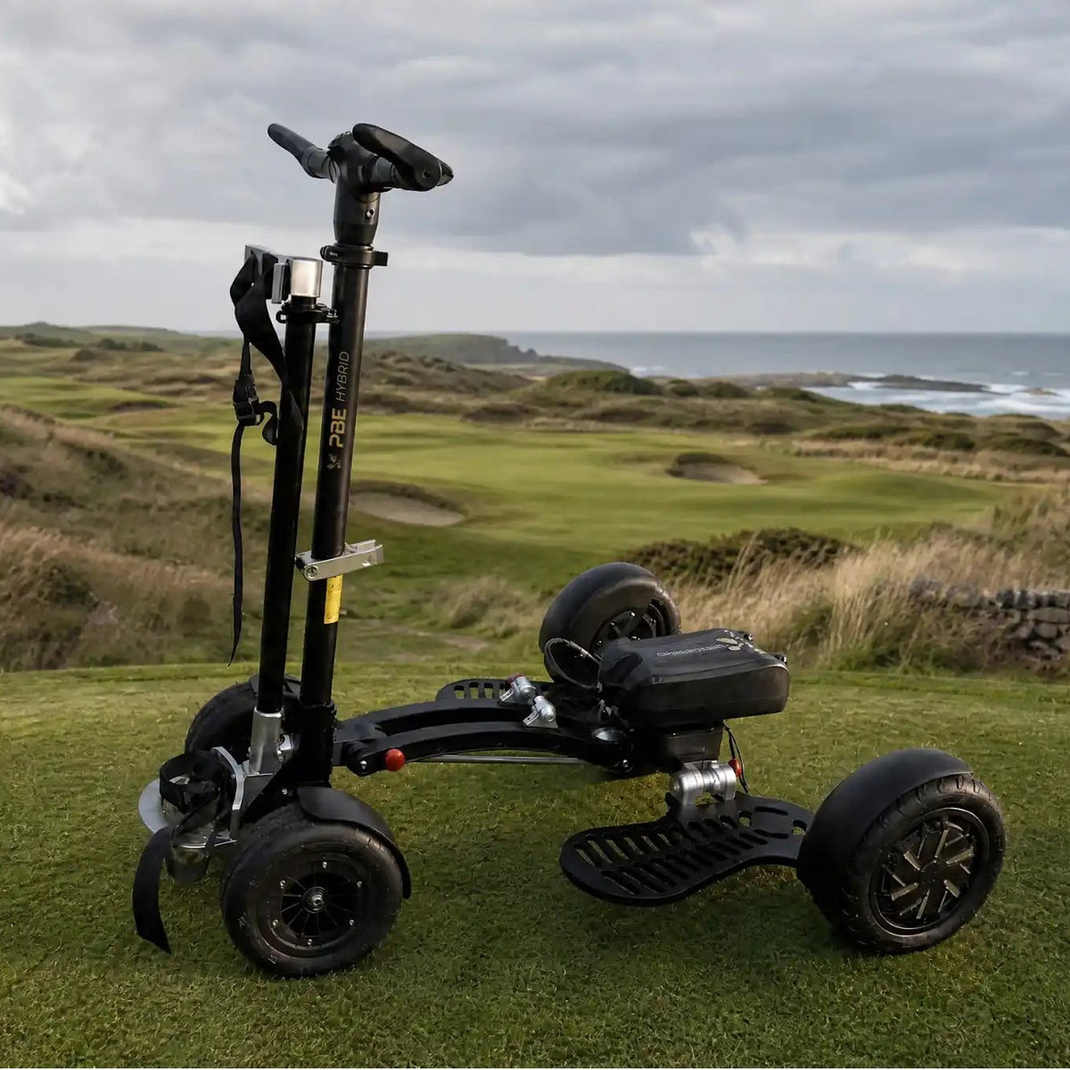 A black three-wheeled golf cart or electric mobility scooter stands on a grassy golf course with rolling green hills, shrubs, and the ocean under a cloudy sky in the background.