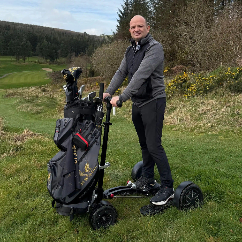 Man standing with a golf trolley and scooter on a grassy area with trees in the background