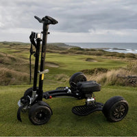 A black three-wheeled golf cart or electric mobility scooter stands on a grassy golf course with rolling green hills, shrubs, and the ocean under a cloudy sky in the background.